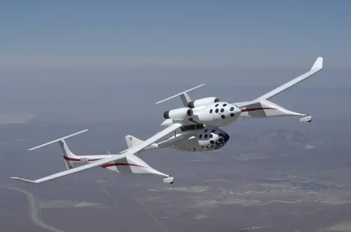 A white aircraft with twin fuselages flies over a desert landscape under a clear sky.