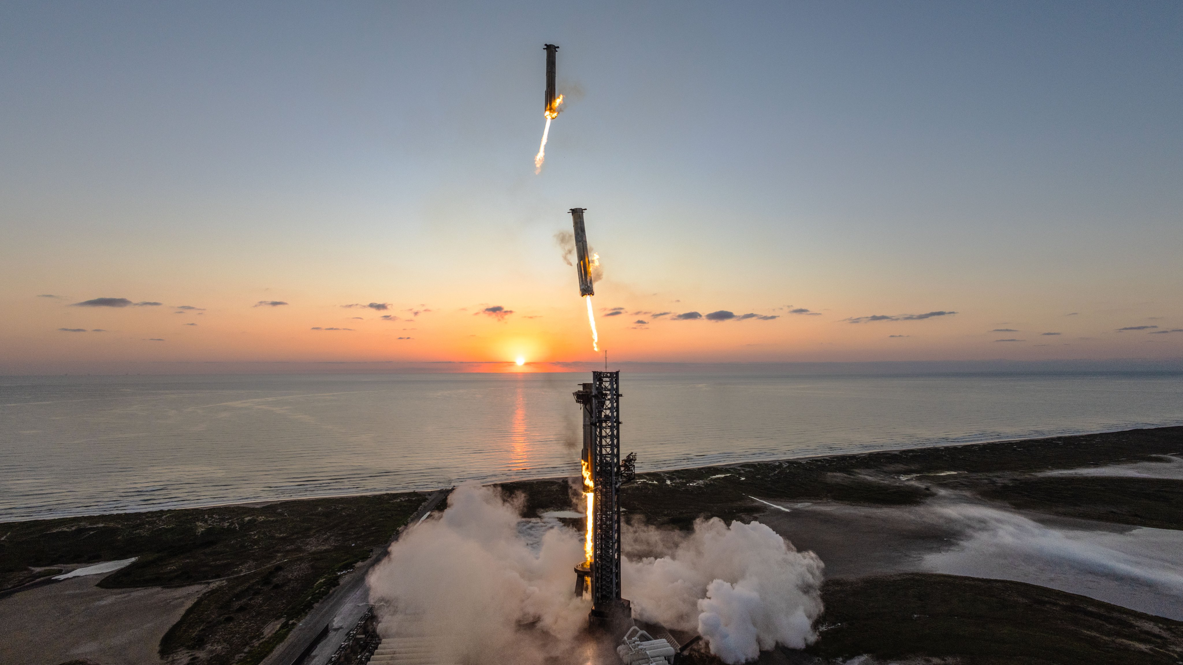 A rocket launches with visible flames and smoke against a sunset over the ocean.