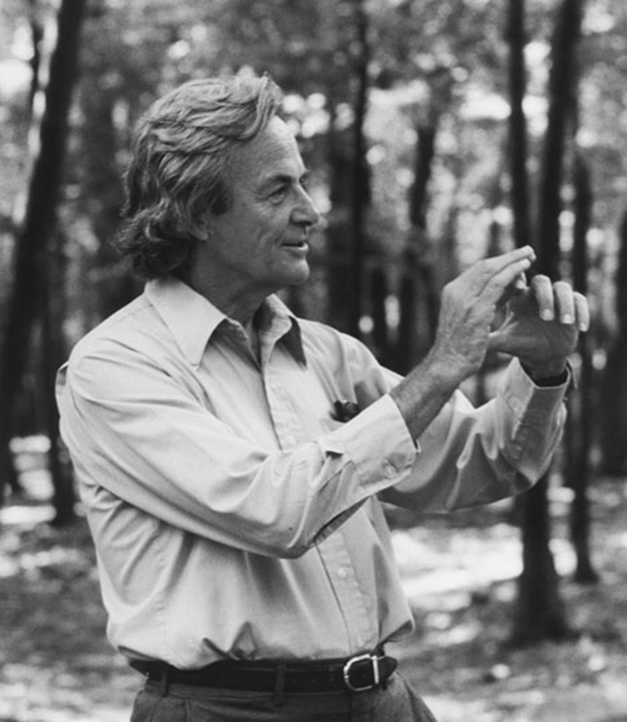 A man in a button-up shirt gestures with his hands while standing in a forested area.