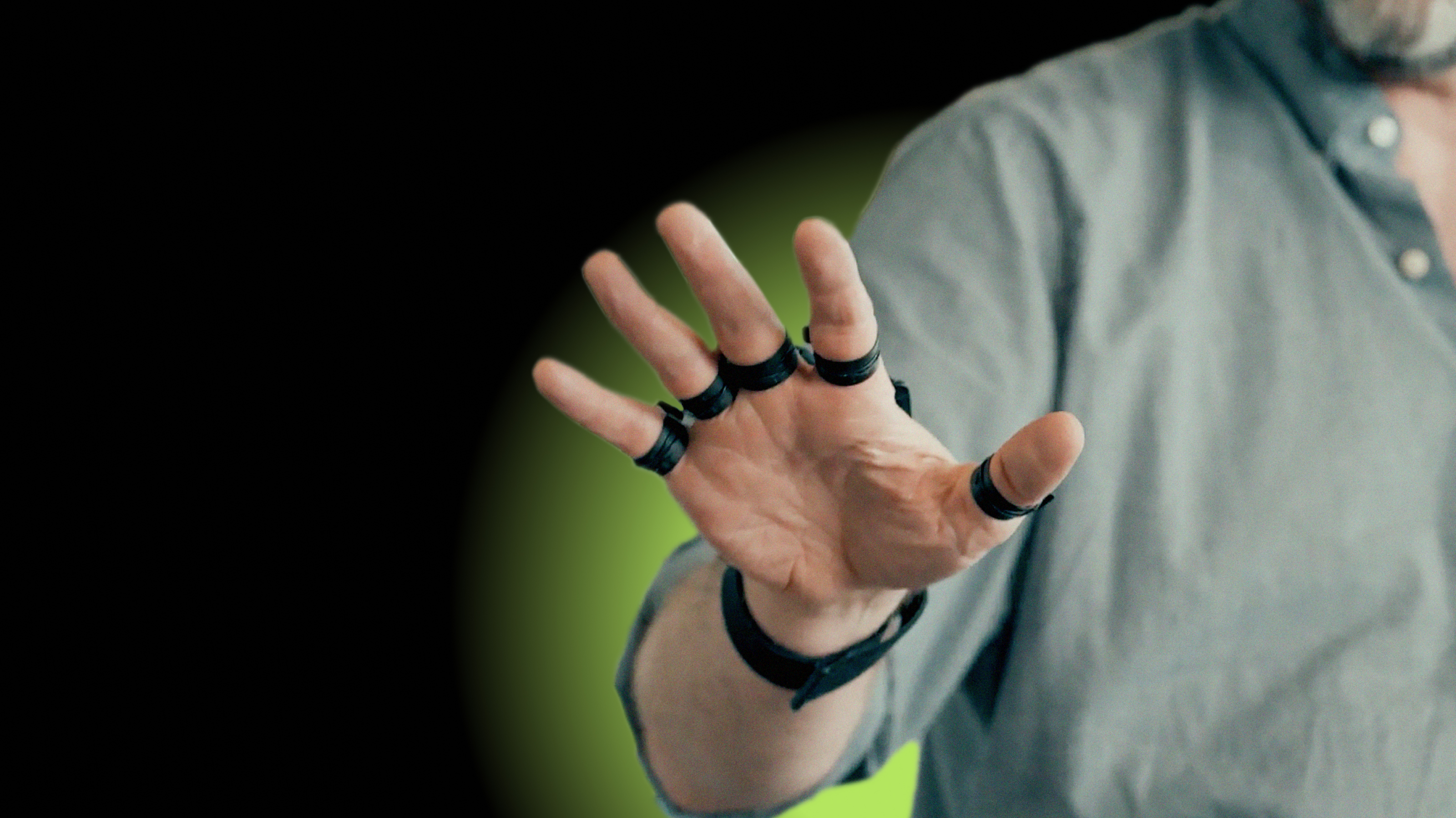 A person wearing a gray shirt is extending their hand with rings on each finger against a dark background with a green glow.
