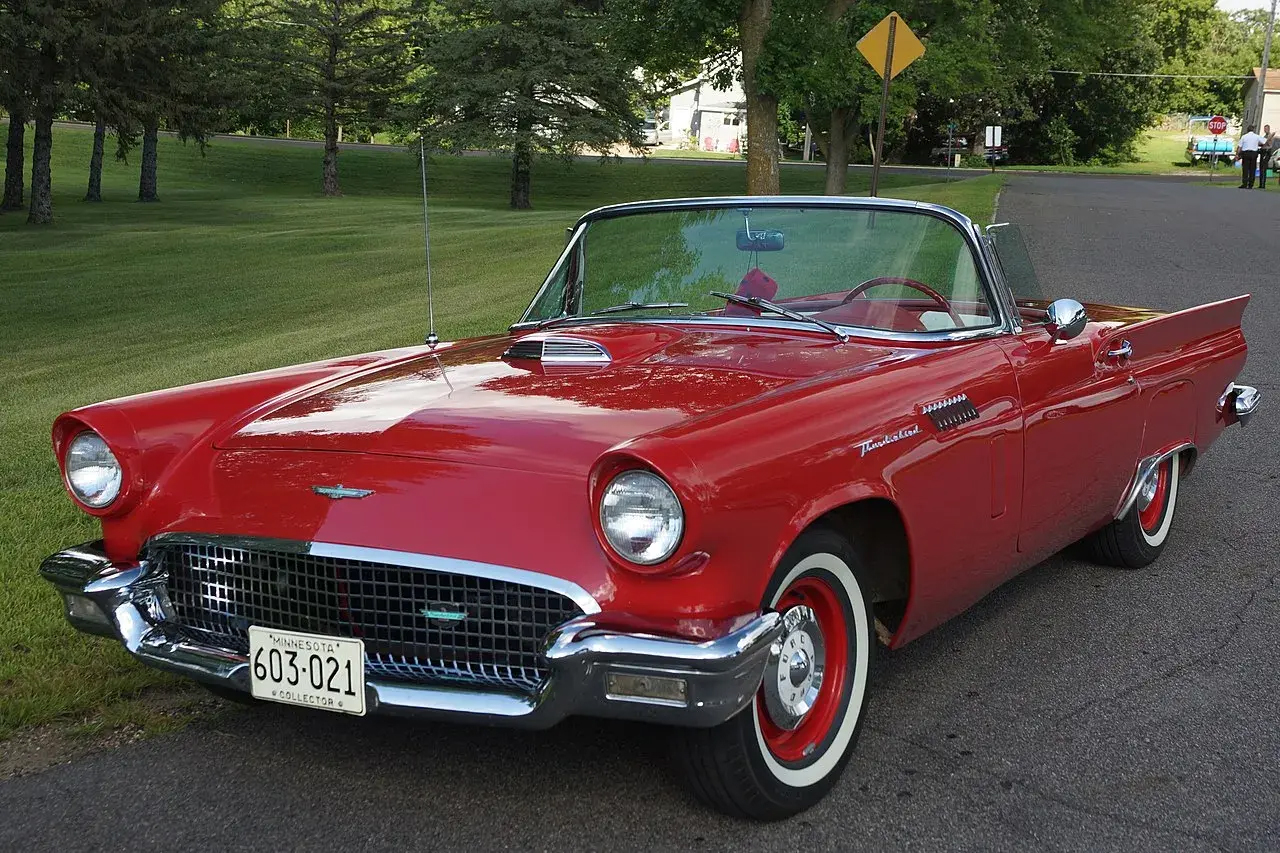 A red vintage convertible car is parked on the side of a suburban road, with trees and grass in the background.