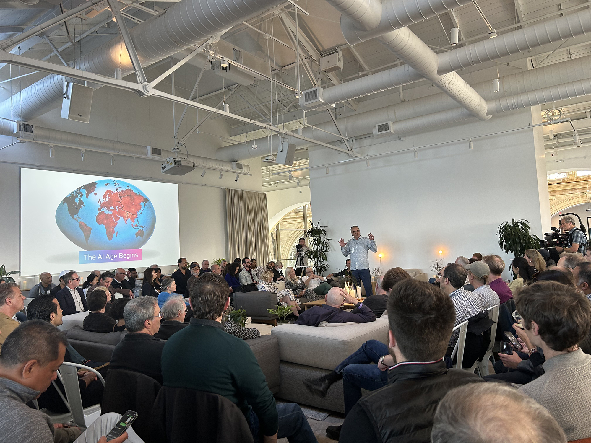 A speaker addresses an engaged audience in a modern, open space. A presentation slide shows a globe with "The AI Age Begins" text.