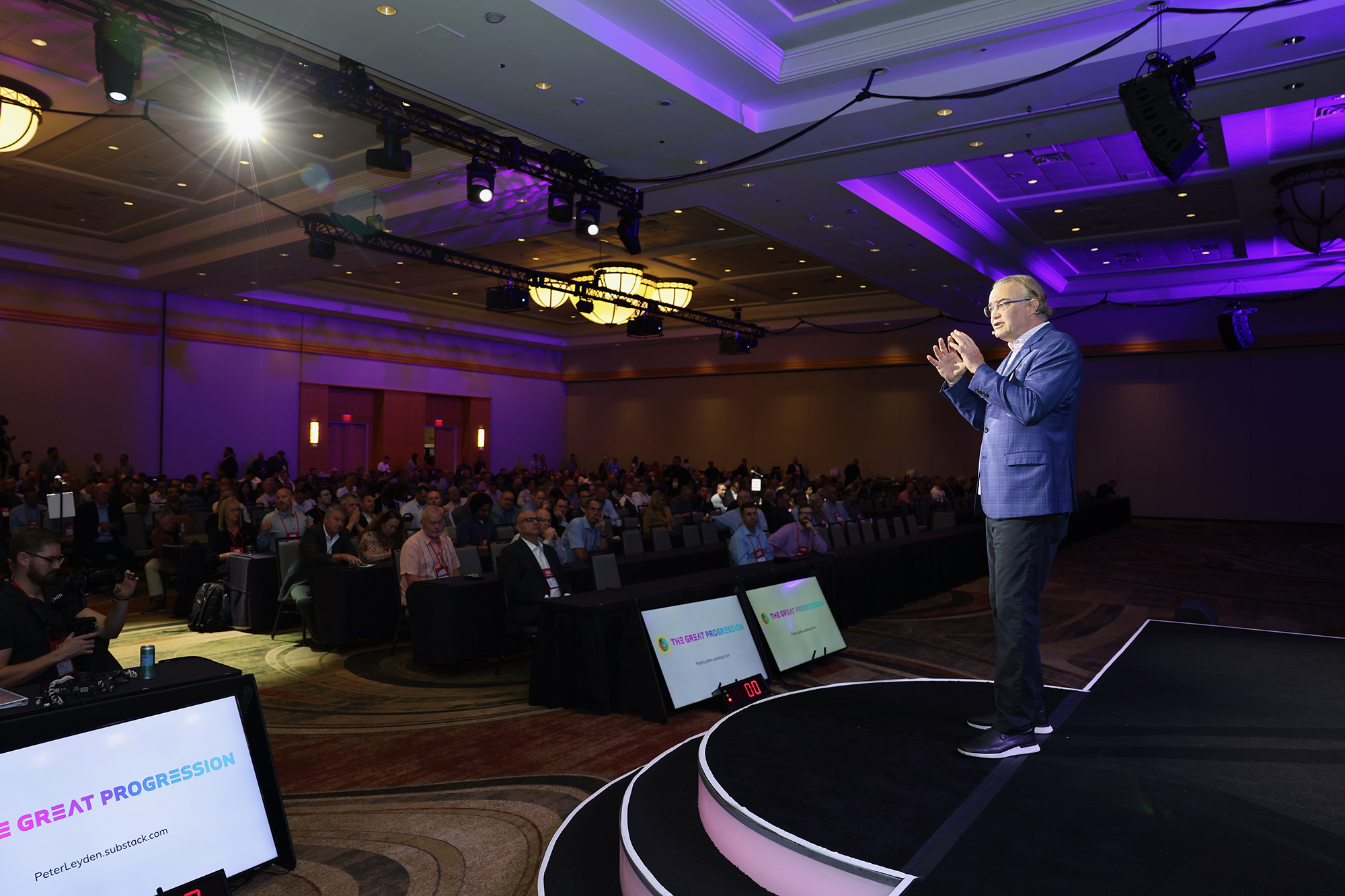 A person speaking on stage to a large audience at a conference, with two screens displaying the words "Great Progression.