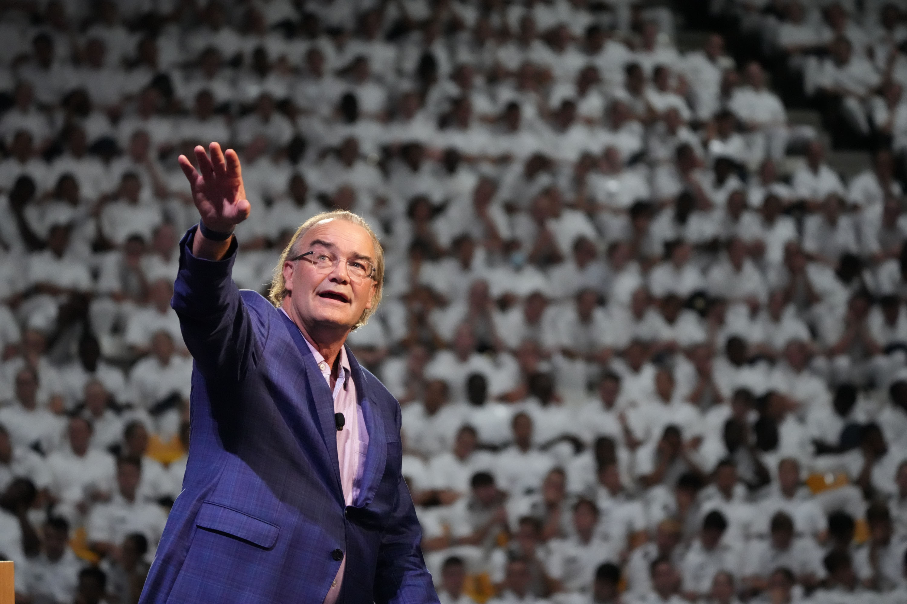 A man in a suit gestures while speaking on stage, with a large audience in white uniforms seated behind him.
