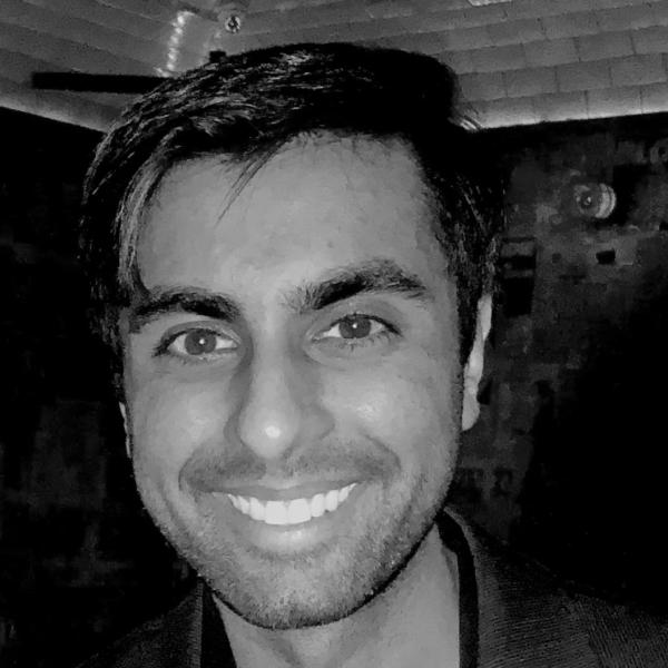 A man with short dark hair and a trimmed beard smiles at the camera in a dimly lit indoor setting.