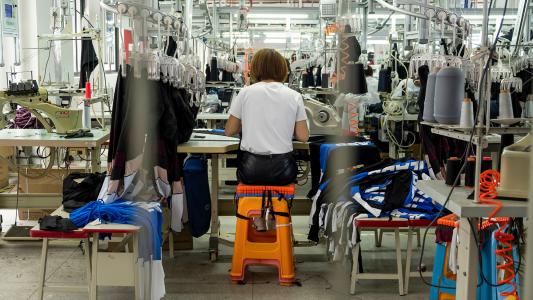 Person sewing at a workstation in a garment factory, surrounded by sewing machines, fabric, and threads.