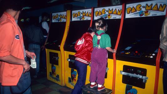 Two children play Pac-Man arcade machines in a dimly lit arcade. One child stands on a stool, while an adult nearby holds cash.