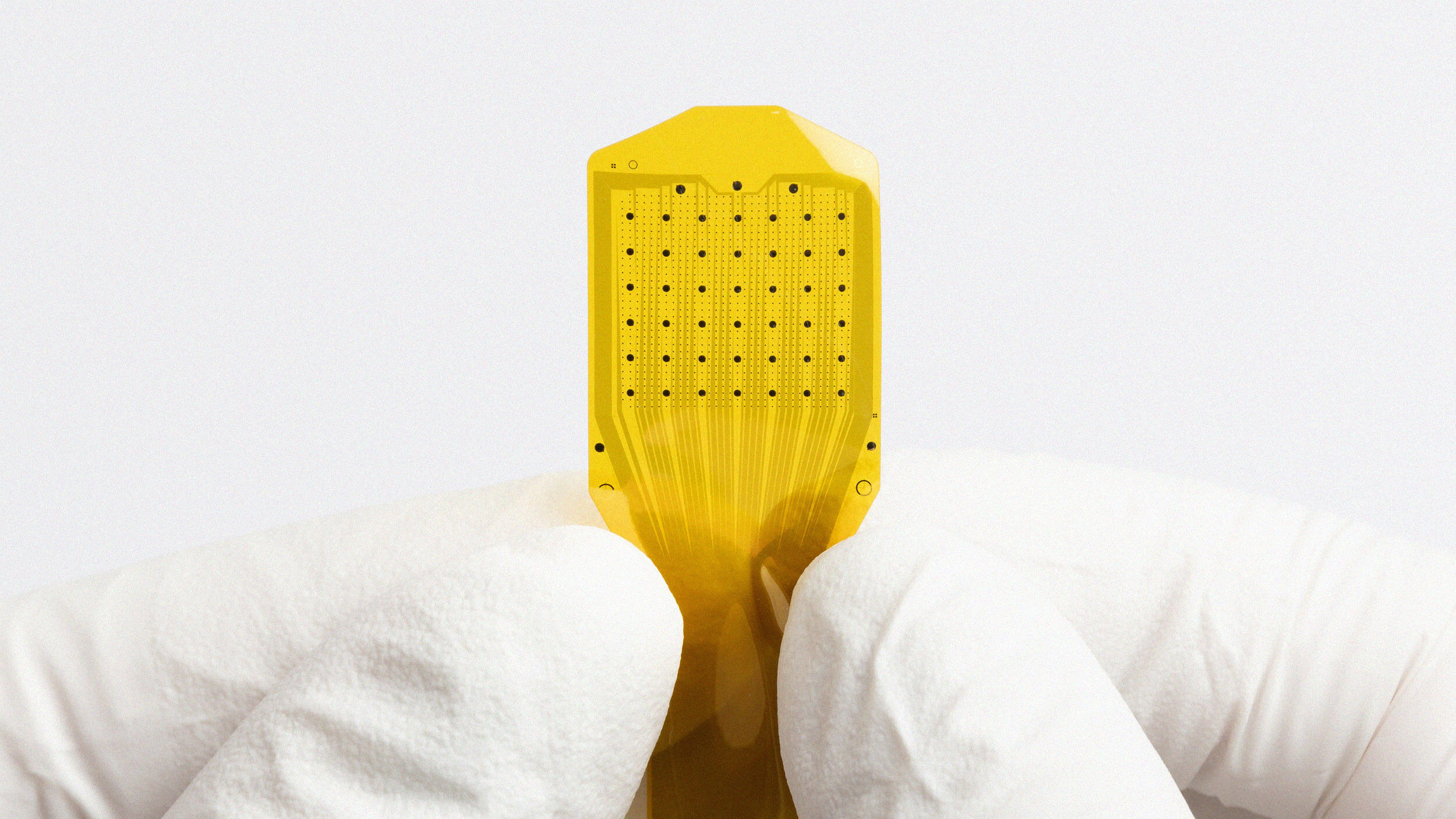 A gloved hand holds a small, yellow electronic microchip with a grid of black dots on its surface against a plain white background.