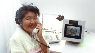 A woman speaks on a phone. A monitor on her desk shows the face of a man.