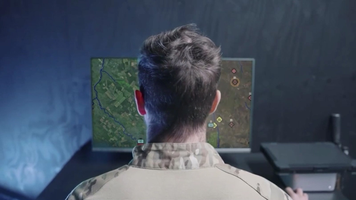 A person in military uniform sits at a desk, looking at a computer monitor displaying a digital map with various symbols and markings.