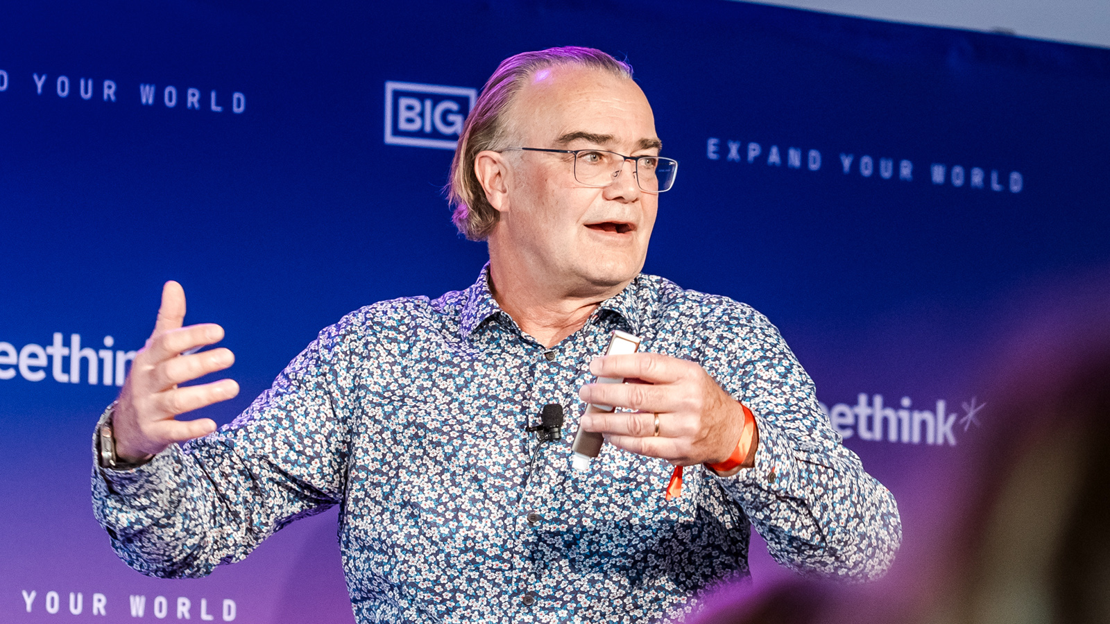 A man with glasses and a patterned shirt gestures while speaking on stage in front of a blue background with white text.
