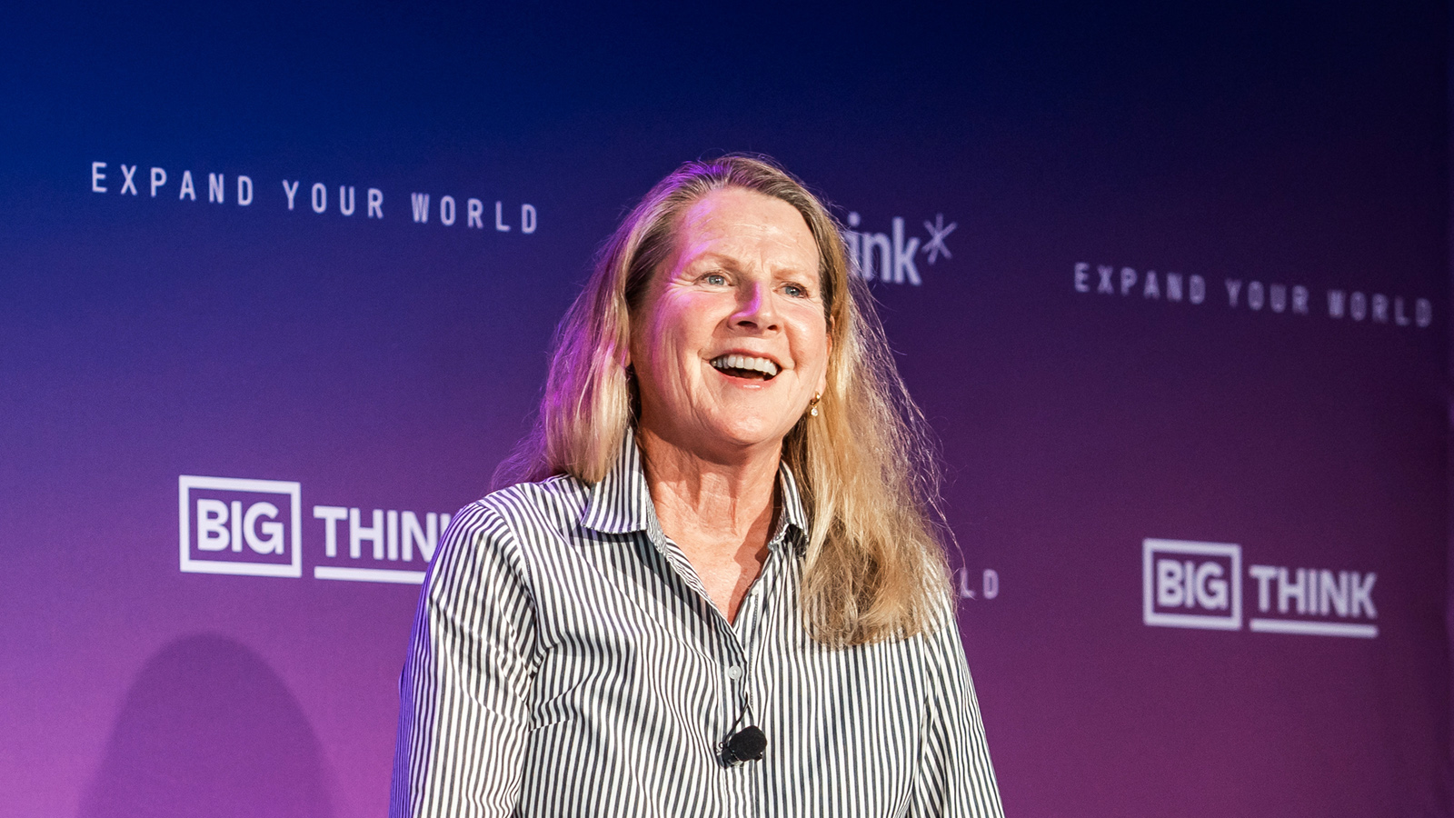A woman with long blond hair wearing a striped shirt smiles while standing in front of a purple backdrop with "BIG THINK" and "EXPAND YOUR WORLD" logos.