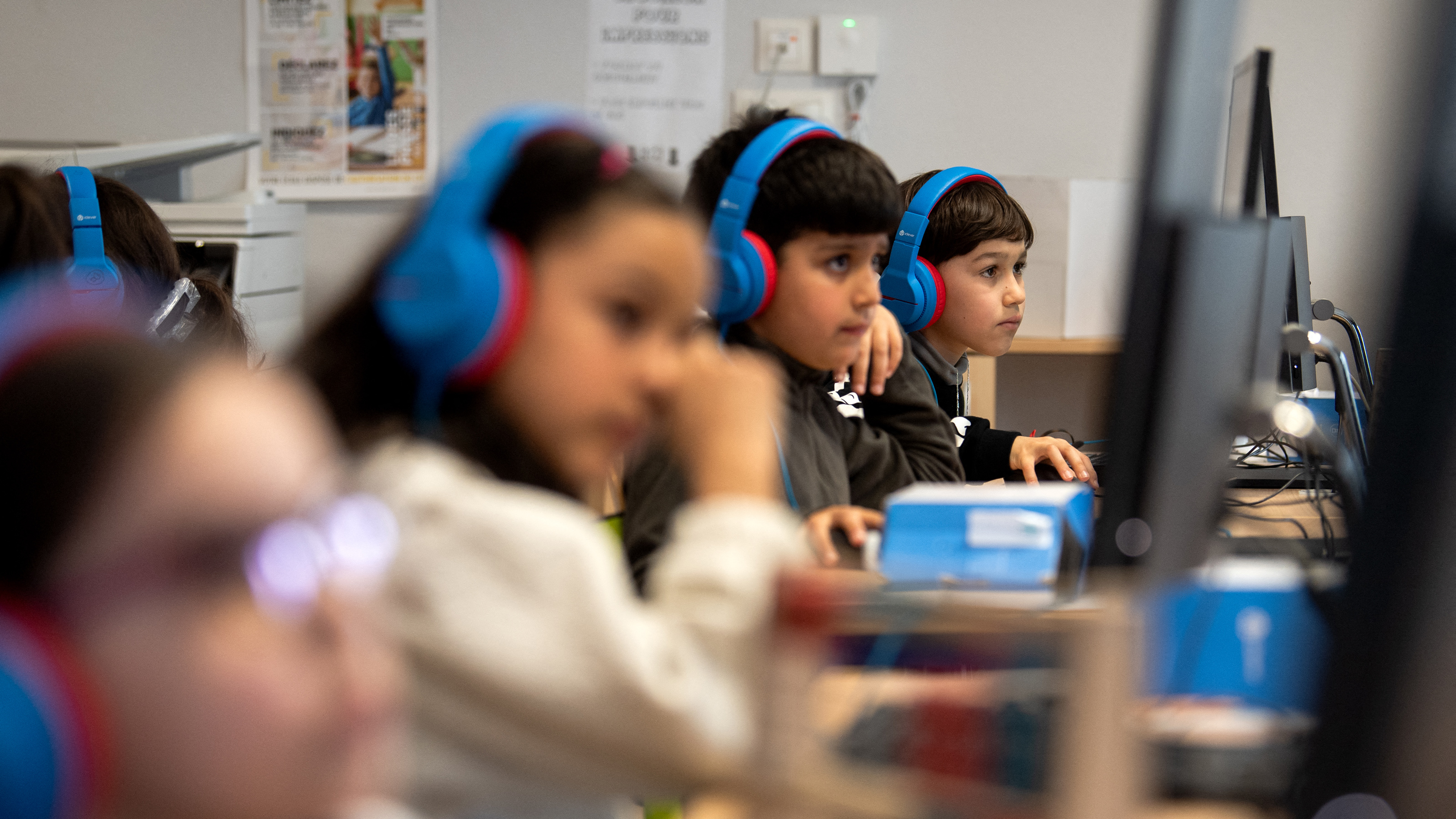 Four children wearing blue headphones use desktop computers in a classroom, focusing on their screens.