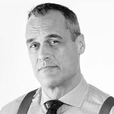 A middle-aged man with short gray hair wearing a striped dress shirt, tie, and suspenders, looking directly at the camera against a plain background.