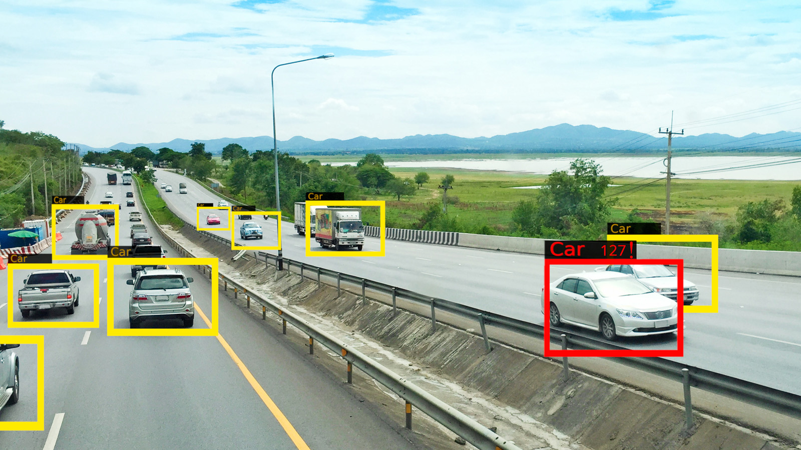 A highway with several cars and trucks labeled in colored boxes, bordered by greenery and a body of water under a cloudy sky.