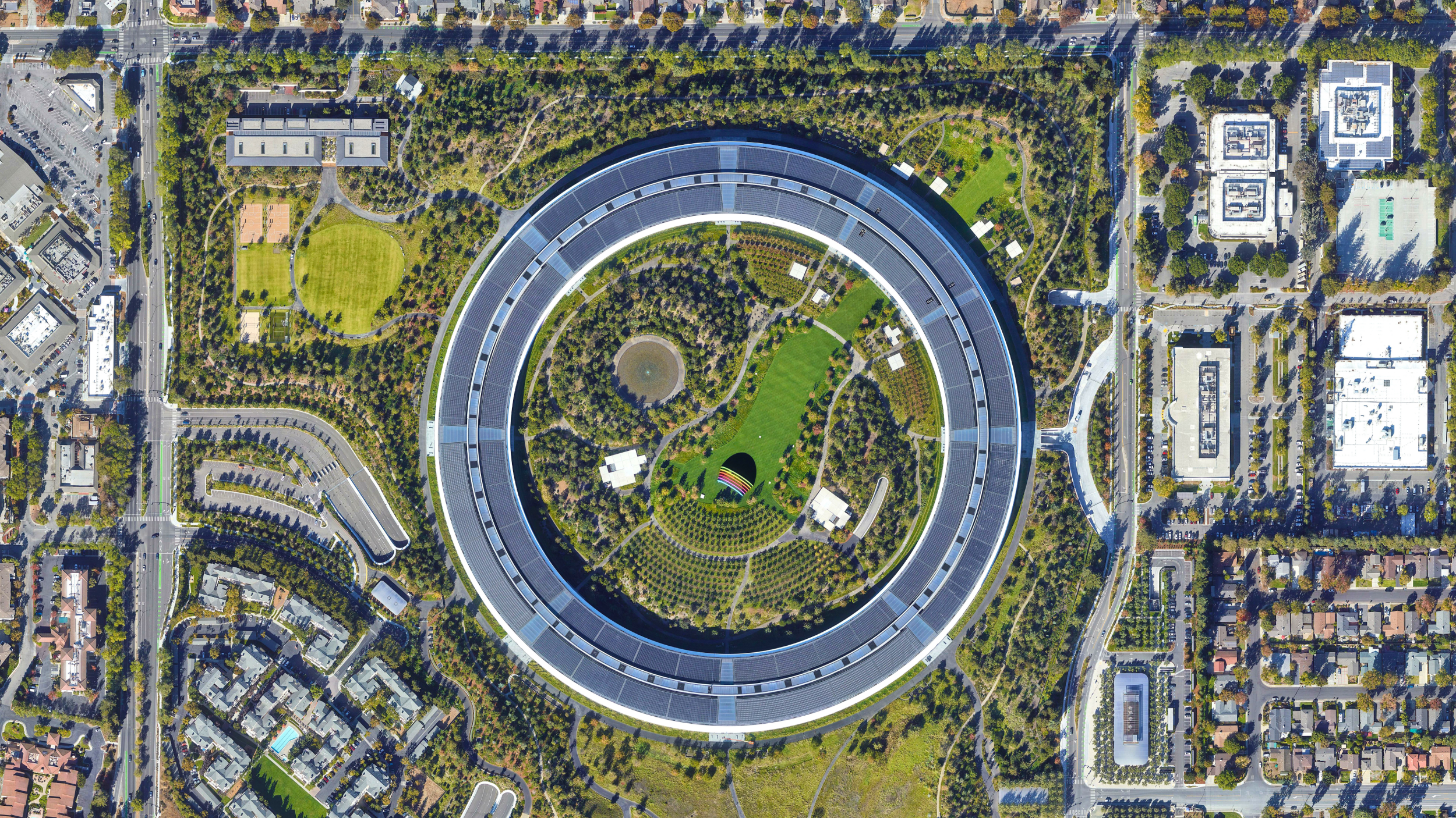 Aerial view of a large circular office building surrounded by trees, green spaces, and nearby roads in an urban area.