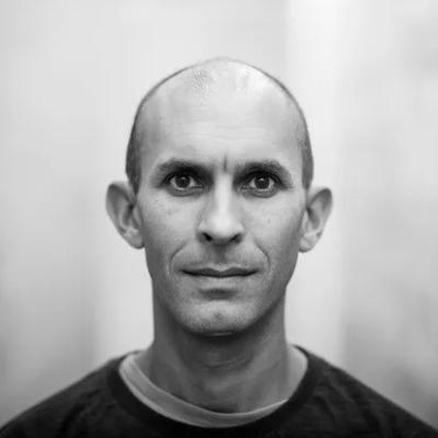 A man with a shaved head and dark eyes stares directly at the camera. The background is blurred and in grayscale.