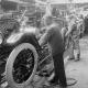 Workers assemble automobiles on a production line in a factory, focusing on tasks near the engines and wheels. Early 20th-century setting with machinery visible in the background.