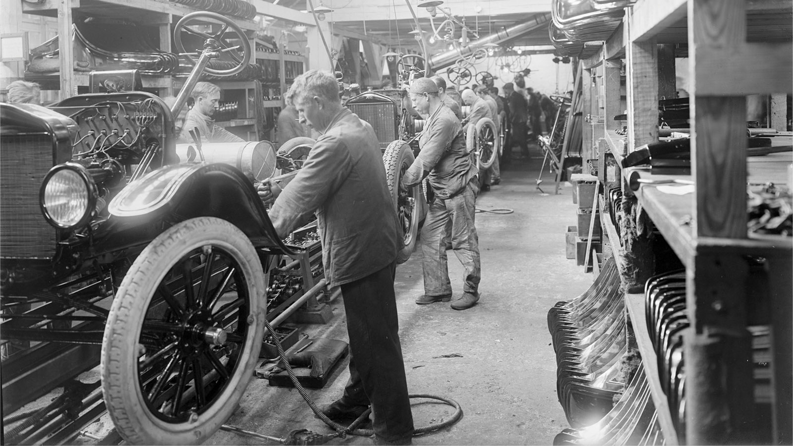 Workers assemble cars in an early 20th-century automotive factory, surrounded by vehicle parts and tools.