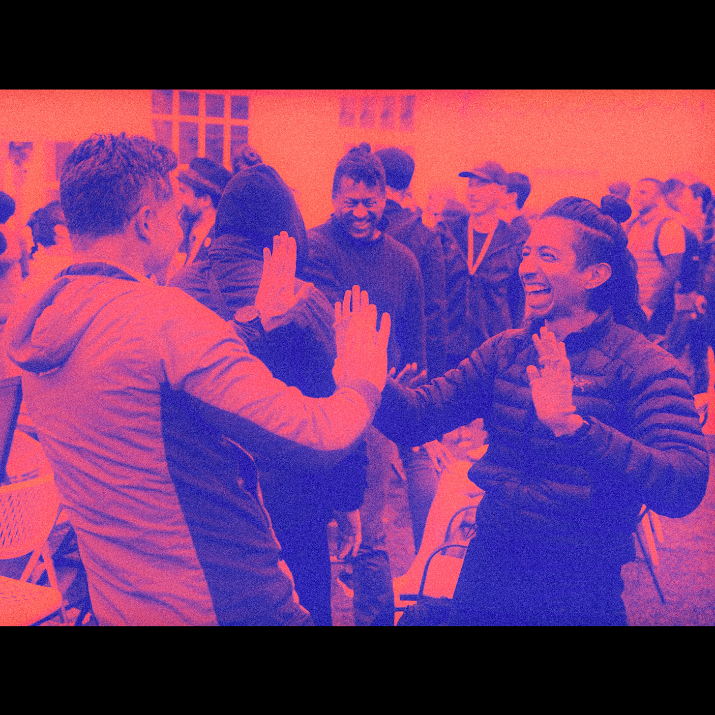 Two people enthusiastically high-five each other in a crowded outdoor setting, surrounded by others who are standing and smiling. Image has a red and blue duotone effect.